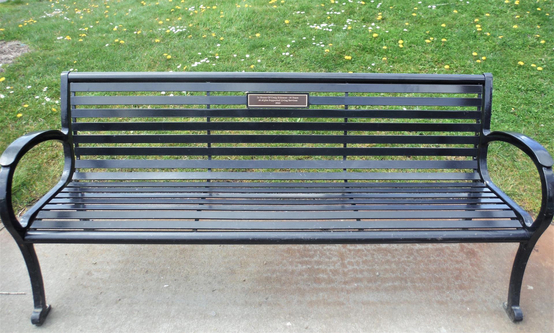 A black, metal bench, approximately six feet long with a commemorative plaque with bronze lettering centered on the back of the bench.