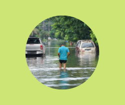 A person walking down a flooded street with two cars in the distance and water reaching their bumpers.