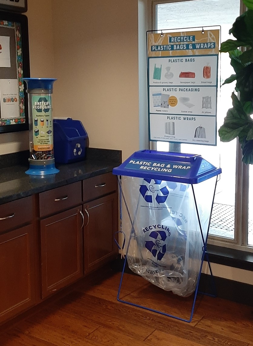 A small battery recycling container on the left and a larger plastic bag recycling station with a large poster on the right.