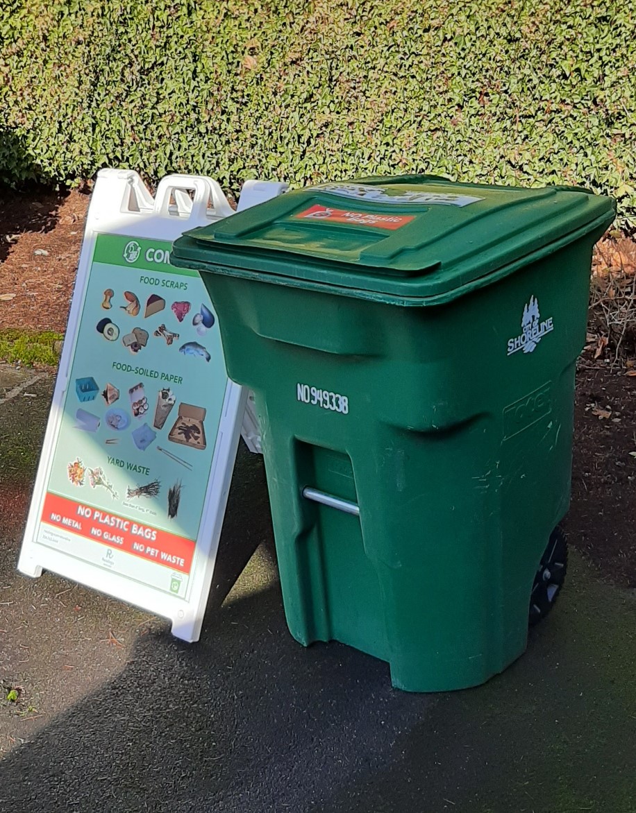 A large green compost cart on the right with a 3-foot tall sign on what can be composted on the left.